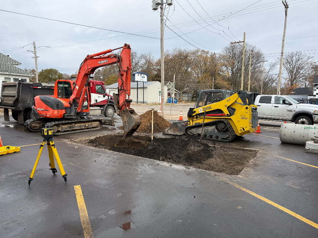 Excavator and skid steer digging a utility trench in a commercial parking lot in Norton Shores, Michigan by Phillip Thomas Excavating (PTE).
